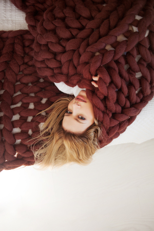 cute young blonde is lying on the bed wrapped in a brown cozy blanket. Warmth and comfort of home, conceptの写真素材