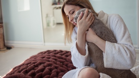 beautiful young blonde girl in a Terry white robe in her apartment hugs her beloved cat. young woman at home playing with a catの写真素材