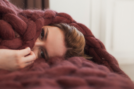 cute young blonde is lying on the bed wrapped in a brown cozy blanket. Warmth and comfort of home, conceptの写真素材