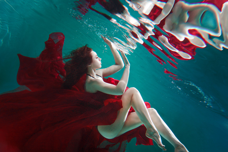 Mystical underwater portrait of a beautiful young woman in a red dress. The girl swims under the surface of the water and her reflection, Immersion conceptの写真素材