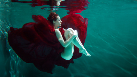 Mystical underwater portrait of a beautiful young woman in a red dress. Girl swimming underwater, fantasy and beauty styleの写真素材