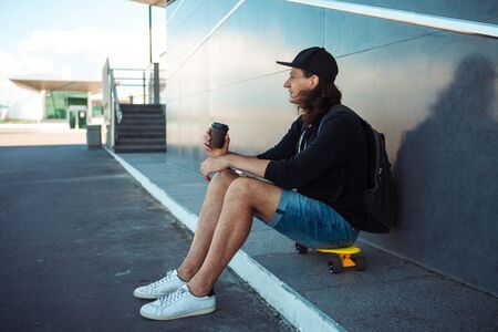 A young man in a baseball cap, with a backpack, and in denim shorts, drinks coffee while sitting on a yellow skateboard on the asphalt, next to a gray granite wall with white stripes. Side view.の写真素材