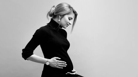 Young beautiful pregnant woman in black clothes hugs her tummy on a neutral background. Black and white. Side view. Copyspace right.の写真素材