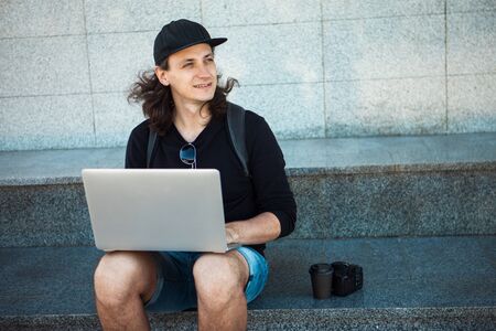 Freelancer working in the open air, concept, young man with long hair sits on the granite steps in the shade, with a laptop. Summer windy day. Copy space. Front viewの写真素材