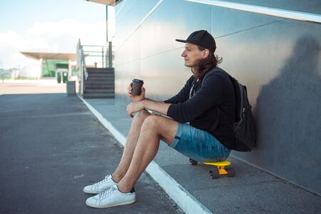 A young man in a baseball cap, with a backpack, and in denim shorts, drinks coffee while sitting on a yellow skateboard on the asphalt, next to a gray granite wall with white stripes. Side view.の写真素材