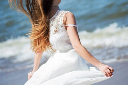 Young charming brunette woman on sea coast. Beautiful girl in a white summer dress. Runs towards the sea, back viewの写真素材
