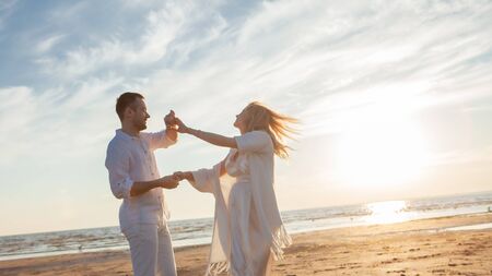 Love, waiting for the baby. Couple, pregnant woman and man, in white flying clothes, walk, hold hands, dance gainst the backdrop of the sunset sea, sandy beach and sky with clouds.の写真素材