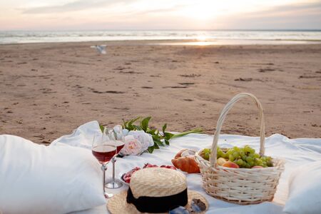 Picnic in the rays of the setting sun on the sand. On the plaid lies a basket of fruit, glasses, flowers and sweet pastries. Nearby is a straw hat.の写真素材