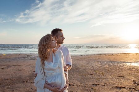 Romantic walk on the beach, waiting for a baby. Happy young couple hugging tummy, spending time by the sea, relaxing, resting, enjoying, against the backdrop of a soft sunset sun.の写真素材