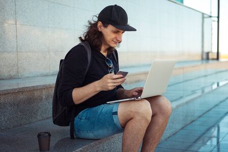 A young brunette man with long hair is sitting on the granite steps in the shade with a laptop on his lap and a smartphone. Summer windy day. Copy space. Side viewの写真素材