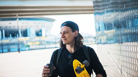 A young man in a baseball cap and denim shorts and with a yellow skate is goes by a shiny metallic reflective wall with urban ornament and a reflection of the bridge. Copy spaceの写真素材