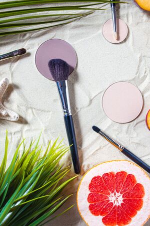 Dry cosmetic decorative product in a round refile lies on the sand, surrounded by bright fruits, corals, and palm leaves. Powder, highlighter, bronzer, blush with a brush. Close-up, top viewの写真素材