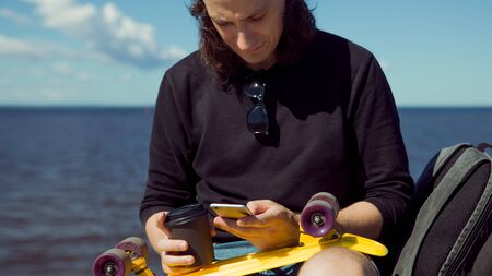 A young man in a baseball cap with a smartphone and a yellow skateboard sits on the parapet of the...