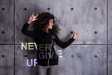 Cheer up! Cheerful young curly woman dancing on a concrete wall background. Multi-colored light inscription, the projection says: Never give up. Copyspace.の写真素材