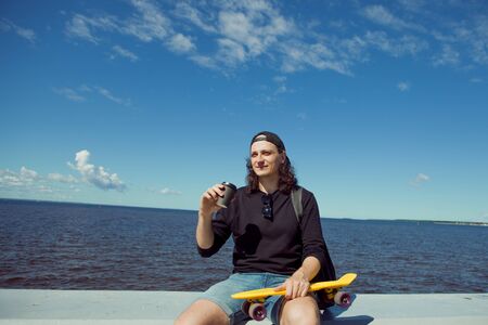 A young man with long hair in a baseball cap, denim shorts, sweatshirt and black glasses is sitting...