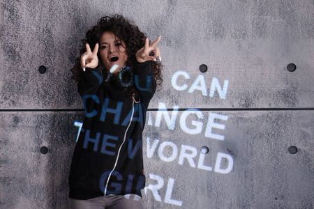 A perky curly girl dances against the background of a gray wall onto which a multi colored motivational inscription is projected. You can change the world, girl. Copyspace.の写真素材