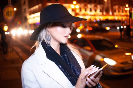 Stylish young woman in an elegant beige coat uses a smartphone standing by the road, on the street...