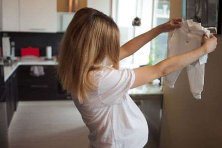 Pregnant happy woman examines small baby clothes with cheerful buttons, holds on outstretched arms. Pretending to try on. Homely atmosphere.の写真素材