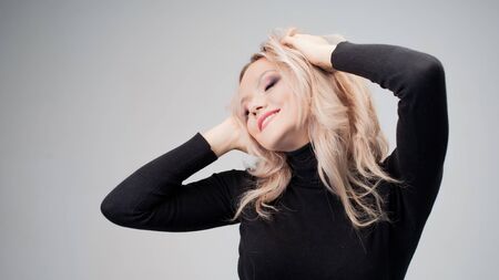 Charming blonde in a black turtleneck with sloppy curls, smiles and touches her hair