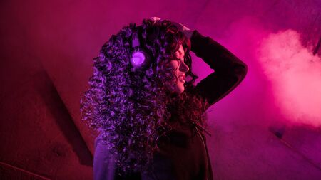 Young beautiful curly woman in big headphones listens to music, sings along, dances against the background of a wall, neon color, magenta.の写真素材
