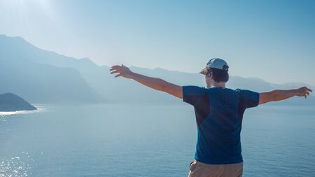 young man stands on top with his arms outstretched, against the sea and the horizon. Back viewの写真素材