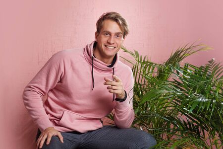 Positive guy in coral hoodie on pink background. Conversation and gestures, pointing the finger at...