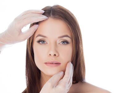 Cosmetology and aesthetic surgery. Portrait of a young attractive woman, clean skin and perfect proportions. Gloved hands touching face, isolated on whiteの写真素材