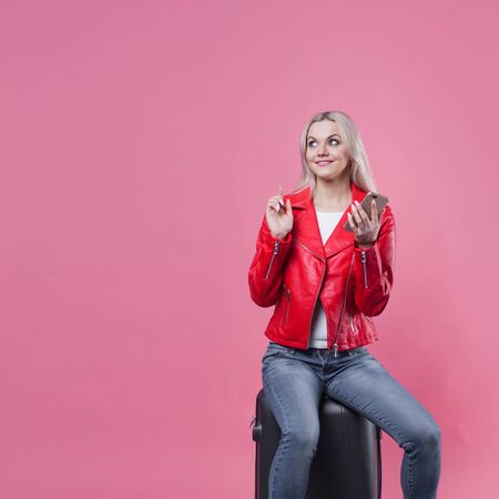 young female tourist in a red jacket and with Luggage on a pink background. Tourist girl ready to fly. Charming blonde sits on a suitcase and checks her smartphoneの写真素材