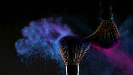 Two Soft cosmetic brushes release a cloud of colored smoke from bright eyeshadow and powder, cosmetics and beauty industry. Bright creative makeup, concept on a black backgroundの写真素材