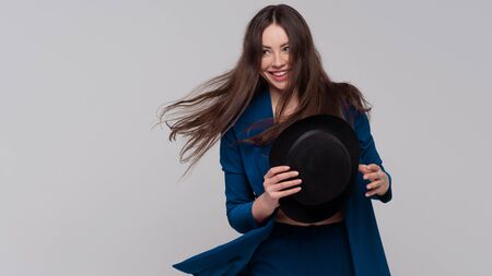 Positive brunette with a hat, trendy and modern girl in a dark blue suit. Studio portrait of a light grey backgroundの写真素材
