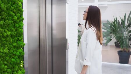 charming young business lady calls the Elevator in the lobby of the business center. Young woman in a white jacket and glasses, stylish brunette at workの写真素材