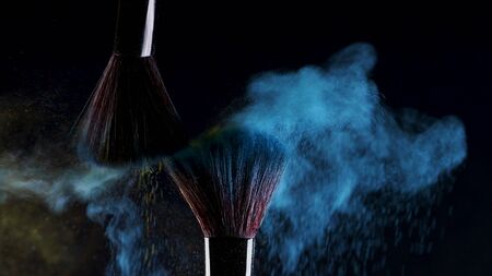 Two Soft cosmetic brushes with blue color release a cloud of colored smoke from bright eyeshadow and powder, cosmetics and beauty industry. Bright creative makeup, concept on a black backgroundの写真素材