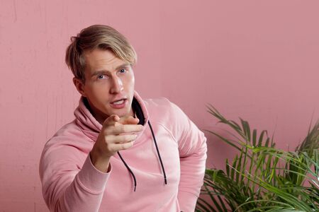 Positive guy in coral hoodie on pink background. Conversation and gestures, pointing the finger at youの写真素材