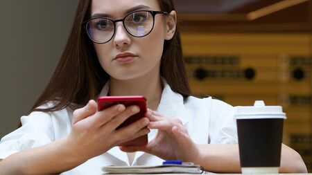 young girl with glasses uses a smartphone, close- up. The student holds in his hands the red phone...
