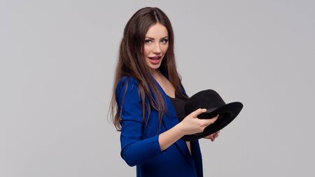 Positive brunette with a hat, trendy and modern girl in a dark blue suit. Studio portrait of a light grey backgroundの写真素材