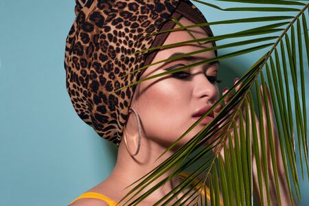 Summer sea and the hot weather, the beach trend. A young beautiful multi-ethnic woman in a leopard turban and yellow swimsuit looks out from behind a palm branch, blue backgroundの写真素材