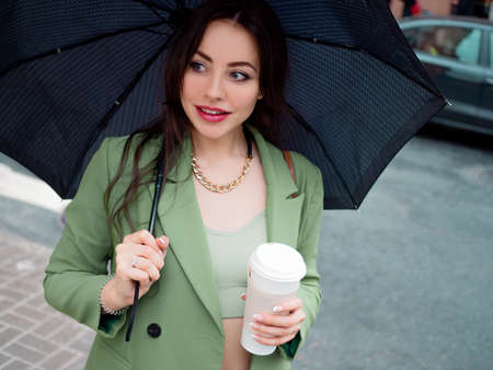 young brunette woman in a green jacket on a walk. Beautiful girl with takeaway coffee with hands...