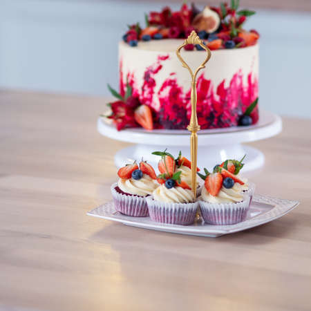Big beautiful red velvet cake, with flowers and berries on top. Delicious sweet muffins with cream, decorated with strawberries and blueberries. Desserts on the kitchen tableの写真素材