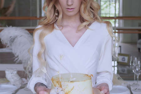 Beautiful elegant bride in a simple white wedding dress with sleeves holding a small wedding cakeの写真素材