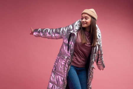 stylish and daring young woman in a silver down jacket dances on a pink background.