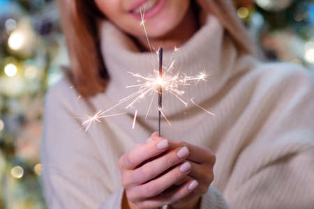 Home new year. A young woman in a cozy white sweater near a sparkling Christmas tree with a Sparkler...