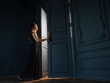 A young woman in a black dress opens a door from which light is pouring.の写真素材