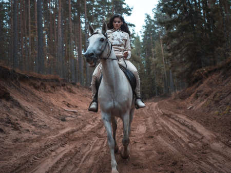 A young beautiful brunette rider in an elegant retro suit riding a white horse in a forest area