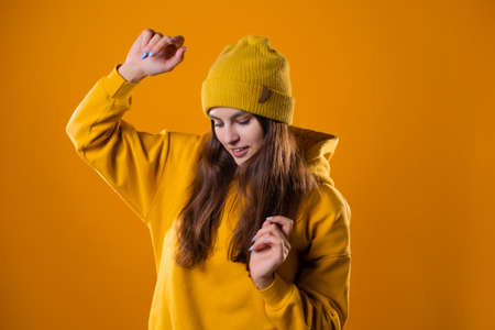 a young cheerful brunette in bright clothes is dancing and having fun. A young woman in a yellow hoodie and a hat on a yellow backgroundの写真素材