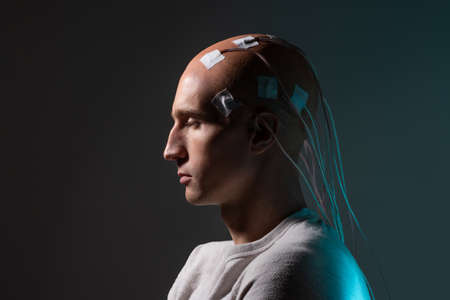 A young man with his head entangled with wires, a portrait in profile, electrical contacts on his bald head, signals to the brain. Neurointerface, future technologies, creative concept.の写真素材
