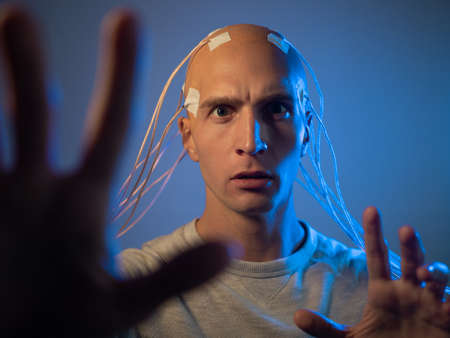 A young man with his head entangled in wires, pulls his hands forward, electrical contacts on his bald head, signals to the brain. Neurointerface, future technologies, creative concept.の写真素材