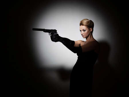 A femme fatale is aiming a silenced pistol, a spy or an undercover agent, a portrait in a spot of lightの写真素材