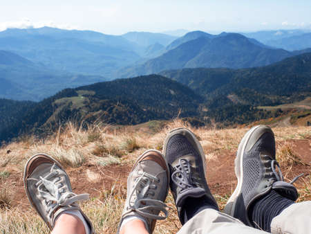 Go to the mountains together. A couple of travelers sat down to relax on the mountain, a first-person view of the mountains, feet in sneakers in the foreground.の写真素材
