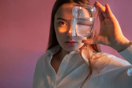 A beautiful young brunette holds a glass of clean water in her hands, the benefits of clean water for beauty and health, concept. Parts of the face are refracted in a glass of waterの写真素材