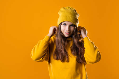 a young cheerful brunette in bright clothes is dancing and having fun. A young woman in a yellow hoodie and a hat on a yellow backgroundの写真素材